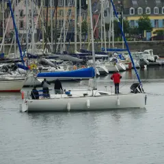 Załoga na jachcie przygotowuje się do manewru, jeden z marynarzy stoi przy sterze, inny pochyla się nad pokładem. W tle port z wieloma żaglówkami i budynkami. Szpring rufowy jest niewidoczny.