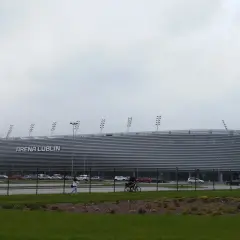 Nowoczesna Arena Lublin z napisem "ARENA LUBLIN" na fasadzie. Obok parking, a na pierwszym planie trawnik. Pojemność obiektu robi wrażenie.