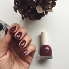 En hand med mörkröda naglar, bredvid en flaska H&M nagellack i samma färg. En torkad blomma i bakgrunden.