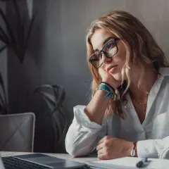 Kobieta w okularach opiera głowę na dłoni, patrząc zamyślona na laptopa.
