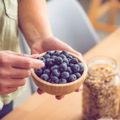 Hände halten eine Schale voller Blaubeeren, reich an Flavonoiden. Daneben stehen Müsli und Bananen – gesunde Flavonoide Lebensmittel für den Start in den Tag.