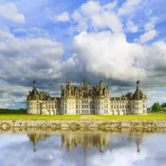 Zamek Chambord nad Loarą odbijający się w wodzie, pod burzliwym, błękitnym niebem.