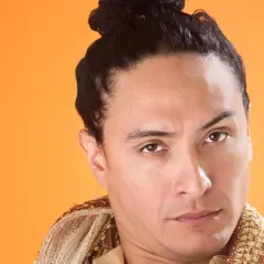 Mann mit dunklem Haar, das zu einem **Samurai-Zopf** gebunden ist, trägt ein reich verziertes Gewand vor orangefarbenem Hintergrund.