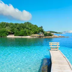Albania - wczasy samolotem all inclusive. Drewniane molo z ławką prowadzi do krystalicznie czystej wody. Zielona wyspa w tle.