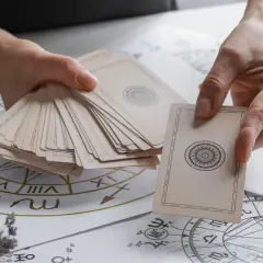 Dłonie trzymają talię kart, przygotowując się do wróżenia. Rozłożone na stole diagramy astrologiczne sugerują, jak stawiać tarota.