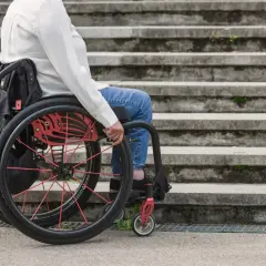 Osoba na wózku inwalidzkim, część grupy inwalidzkiej, siedzi przed kamiennymi schodami.