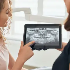 Pacjentka z kręconymi włosami ogląda z dentystką panoramiczny rentgen stomatologiczny na tablecie.