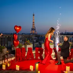 Proposition romantique à Paris, avec la Tour Eiffel en arrière-plan. Un homme s'agenouille, une bague à la main, devant sa compagne. Des roses rouges forment le mot "MARRY ME". Une violoniste joue, des feux d'artifice illuminent la scène. Un moment dig...