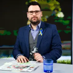 Michał Szajder, wiek nieznany, w garniturze z przypinką w barwach Ukrainy, siedzi przy stole z gazetami i szklanką wody.