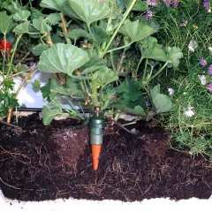 Gröna växter med röda blommor och lila blommor i en låda. Tre gröna droppbevattningskottar är nedstuckna i jorden, kopplade till ett självvattningssystem.