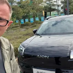 Mężczyzna w okularach pozuje do selfie obok czarnego SUV-a marki AION, jednej z nowych marek samochodów.