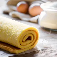 Rotolo di pasta biscotto per torte a strati, pronto per essere farcito. Sullo sfondo, uova e zucchero.