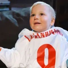 Uśmiechnięty chłopiec z zespołem Angelmana w białej bluzie z czerwonym napisem i cyfrą 9.