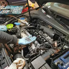Mechanik w niebieskich rękawiczkach trzyma świece żarowe do wymiany. W tle skrzynka z narzędziami i silnik samochodu.