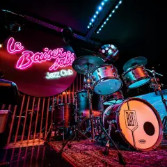 Batterie de jazz dans un bar parisien, éclairé par des néons roses et bleus. Ambiance feutrée pour une soirée musicale.