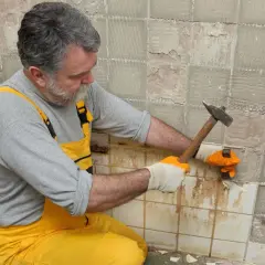 Homme en salopette jaune en train d'enlever du carrelage mural avec un marteau et un burin.