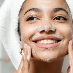 Mujer sonriente con espuma en el rostro, disfrutando de su rutina de doble limpieza facial.