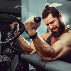 Mężczyzna z brodą i kitką ćwiczy biceps na maszynie. Widać jego napięte mięśnie i determinację. To idealne miejsce, by budować **mutant mass sklad**.