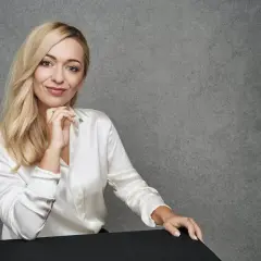 Blondynka w białej satynowej koszuli podczas sesji modowej, z dłonią pod brodą, uśmiecha się do obiektywu.