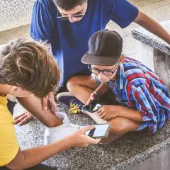 Trzech chłopców pochylonych nad telefonami. Jeden z nich pokazuje coś na ekranie, być może treści pełne hejtu, które mogą wpływać na ich samopoczucie.