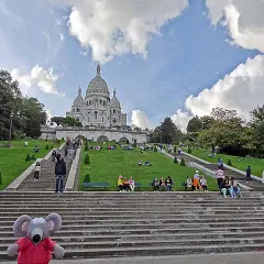 Un adorable raton laveur en pull rouge pose sur les marches menant au Sacré-Cœur, où des visiteurs profitent du soleil sur l'herbe verdoyante.