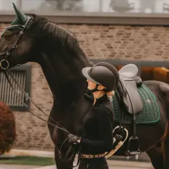 Eleganckie nauszniki zdobią głowę ciemnego konia, gotowego do jazdy. Obok stoi jeźdźczyni w kasku.