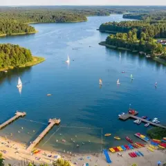 Malownicze jezioro Kałębie z żaglówkami, kajakami i plażą pełną wypoczywających ludzi.