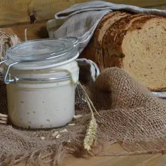 Un barattolo di lievito madre attivo, con una fetta di pane rustico sullo sfondo. Ingredienti naturali per un pane fragrante.