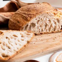 Un delicioso pan de masa madre, con su corteza dorada y miga aireada, reposa sobre una tabla de madera junto a unas rebanadas.