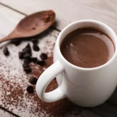 Tazza di cioccolata calda con cacao amaro, gocce di cioccolato e un cucchiaio di legno su un tavolo di legno.