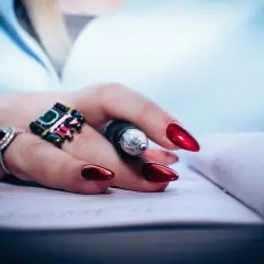 Dłoń z błyszczącymi, czerwonymi paznokciami i pierścionkami trzyma długopis nad notatnikiem. Dbałość o detale, jak witaminy na paznokcie, podkreśla styl.