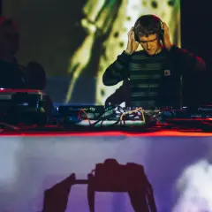 DJ w słuchawkach przy konsoli Pioneer DJ, w tle inny DJ i projekcja.