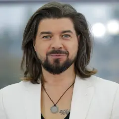 Tomasz Szczepanik w białej marynarce, z długimi włosami i brodą, patrzy w obiektyw.