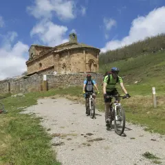 Ciclistas exploran el románico palentino, pedaleando hacia una iglesia de piedra bajo un cielo azul con nubes.