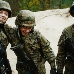Trzech uśmiechniętych żołnierzy wojsk obrony terytorialnej w mundurach i hełmach, z karabinami, na tle piasku i drzew.