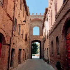 Un vicolo antico con edifici in mattoni rossi e un arco monumentale che conduce a un giardino. Sembra un buon convento.
