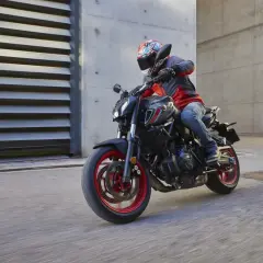Motociclista con casco y chaqueta roja y gris conduce una Yamaha MT-07 negra con detalles rojos por una calle urbana.