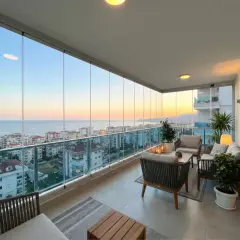 Terraza moderna con cortinas de cristal, muebles de exterior y vistas al mar al atardecer.