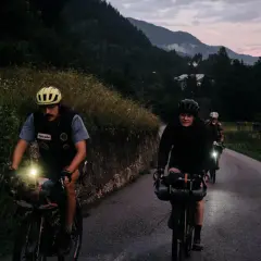 Rowerzyści w kaskach jadą nocą, oświetlając drogę lampkami. Widać, jak działa dynamo, generując prąd do świateł.
