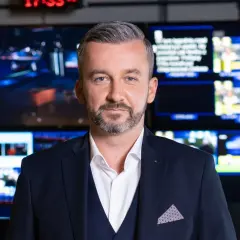 Krzysztof Skórzyński, elegancki mężczyzna w garniturze, stoi na tle ekranów. Ile ma lat? Tego nie wiemy, ale wygląda na doświadczonego dziennikarza.