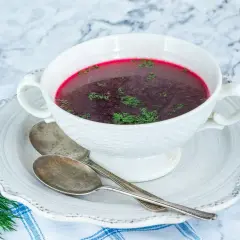 Aromatic barszcz czerwony domowy, ozdobiony świeżym koperkiem, czeka w białej miseczce na talerzyku z dwoma łyżkami.