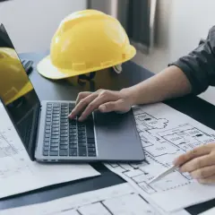 Inżynier analizuje plany, obliczając powierzchnię netto budynku. Na biurku leży żółty kask i laptop.