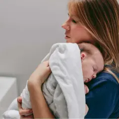 Kobieta tuli śpiące niemowlę. Dziecko ma na sobie biały kocyk, a jego główka spoczywa na ramieniu mamy. Może to być moment ukojenia po czkawce u niemowlaka.