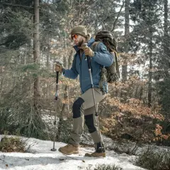 Mężczyzna w zimowej kurtce i czapce idzie przez las, wspierając się na kijach trekkingowych. W tle drzewa i śnieg.