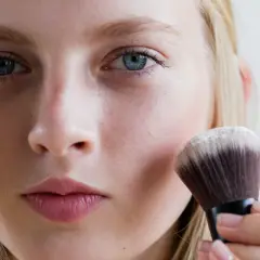 Blondynka nakłada puder transparentny pędzlem. To sekret naturalnego wykończenia makijażu.