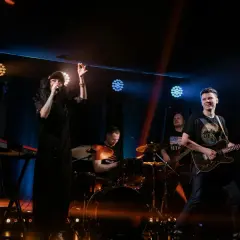 Energiczny zz trio rockowe na scenie. Wokalistka w czarnej sukience, gitarzysta, basista, perkusista i klawiszowiec tworzą dynamiczny zespół.