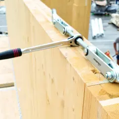 Un outil de serrage métallique, faisant partie d'une liste d'outils de charpentier, est utilisé sur de grosses poutres en bois.