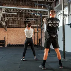 Para osiągnięcia dobrych wyników w crossfit, jak często trenować? Para ćwiczy w siłowni, kobieta z kettlebell, mężczyzna w koszulce "It's Going Down".