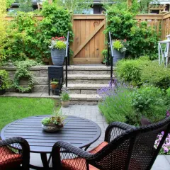 Ein kleiner Garten mit Sitzgruppe, Stufen und Holztor. Üppige Pflanzen und Blumen schaffen eine grüne Oase.