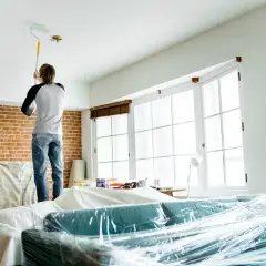 Mężczyzna maluje sufit na biało. Zastanawia się, jaka farba na sufit będzie najlepsza, by odświeżyć wnętrze. Meble zabezpieczone folią.